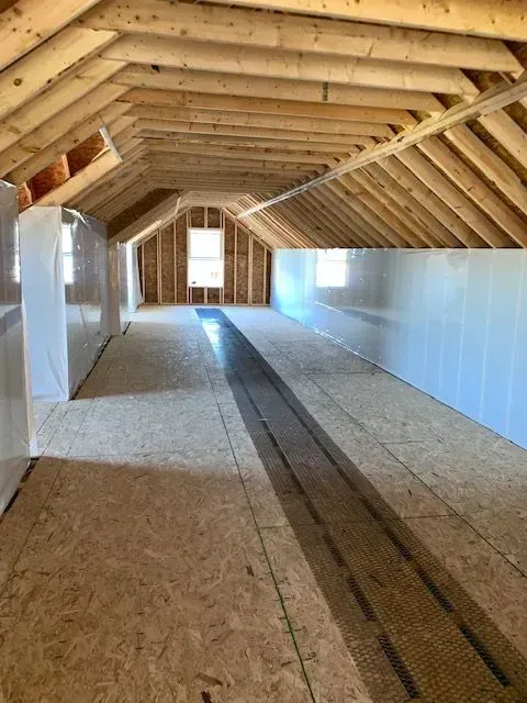 Interior of an attic with exposed wooden beams, plywood floor, and a small window.