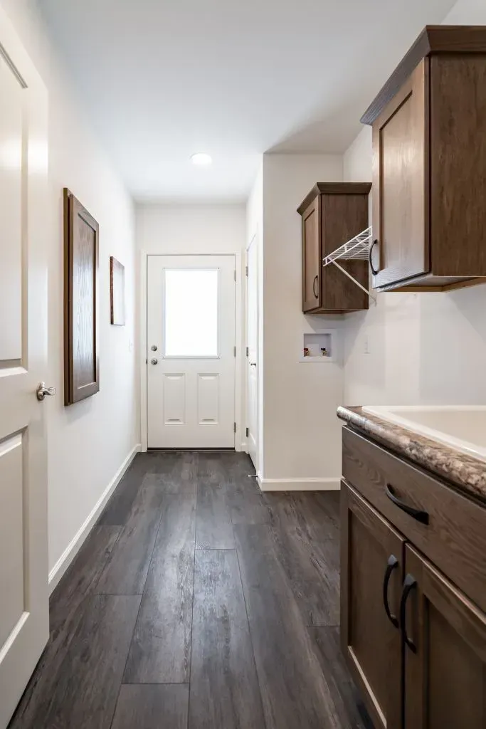 Laundry room with dark wood flooring, white walls, and brown cabinets. Door at the end.