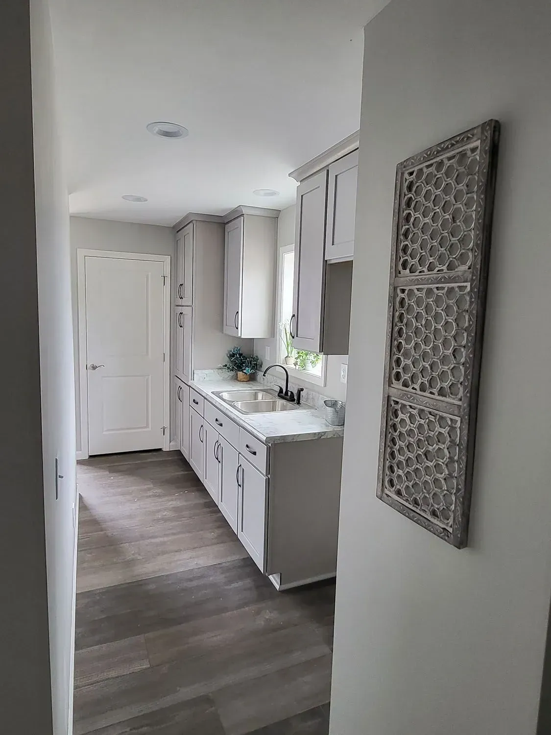 Hallway leading to a light-colored kitchen with gray cabinets, a sink, and a decorative wall panel.