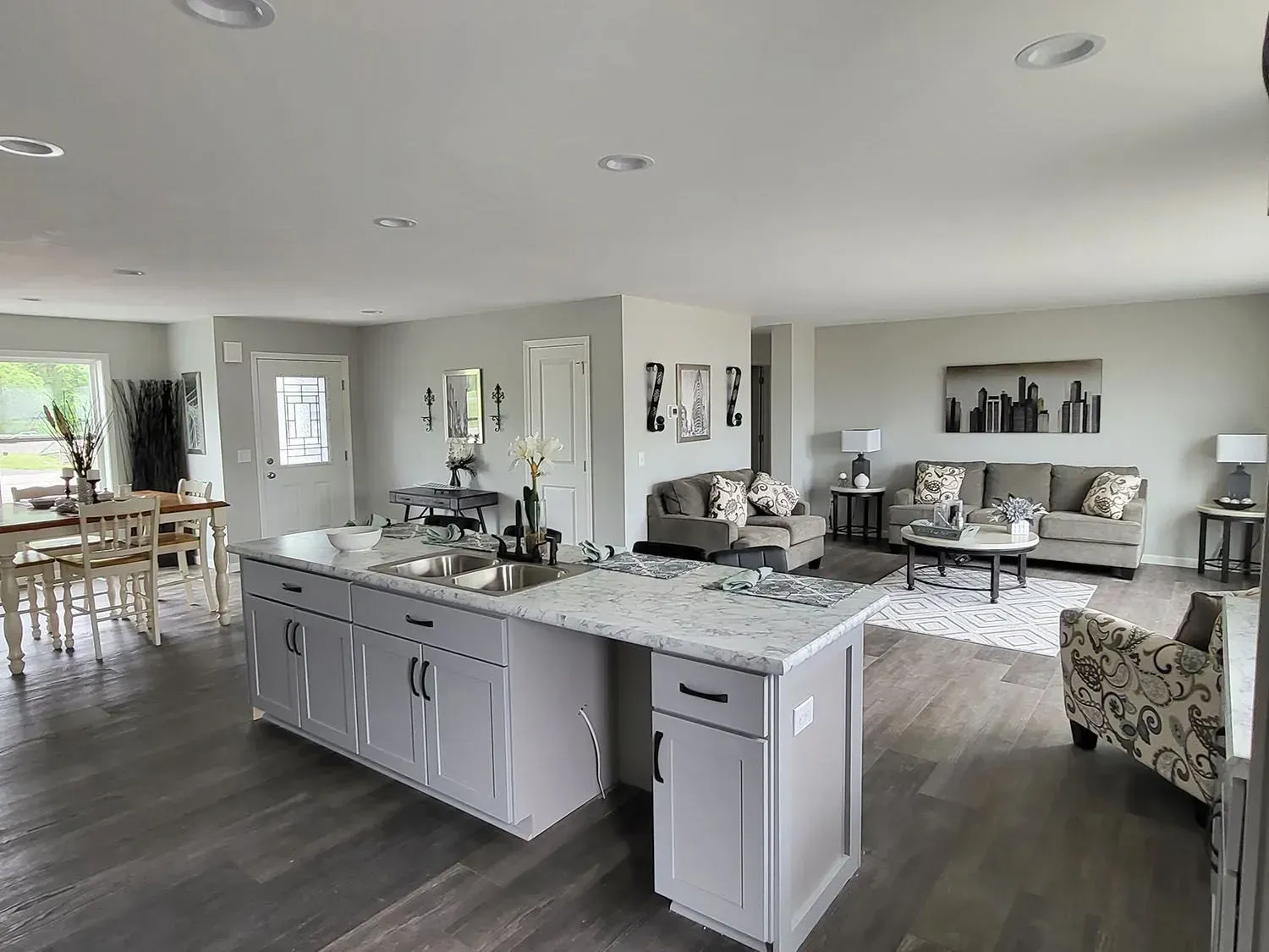 Open-concept living space with kitchen island, dining table, and living room seating. Gray walls and flooring.