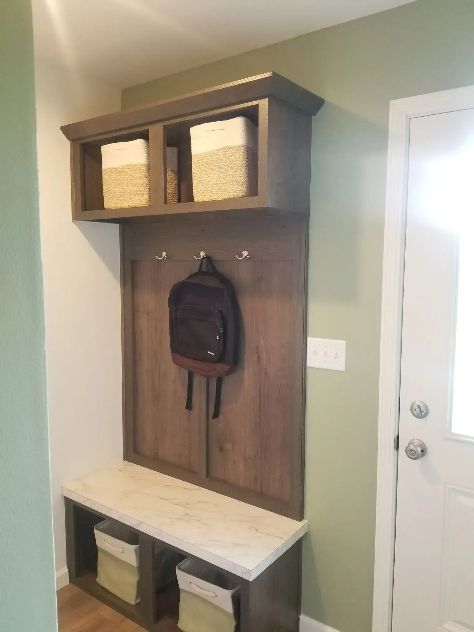 Wooden entryway storage with bench, hooks, and overhead cubbies holding baskets; backpack hangs.