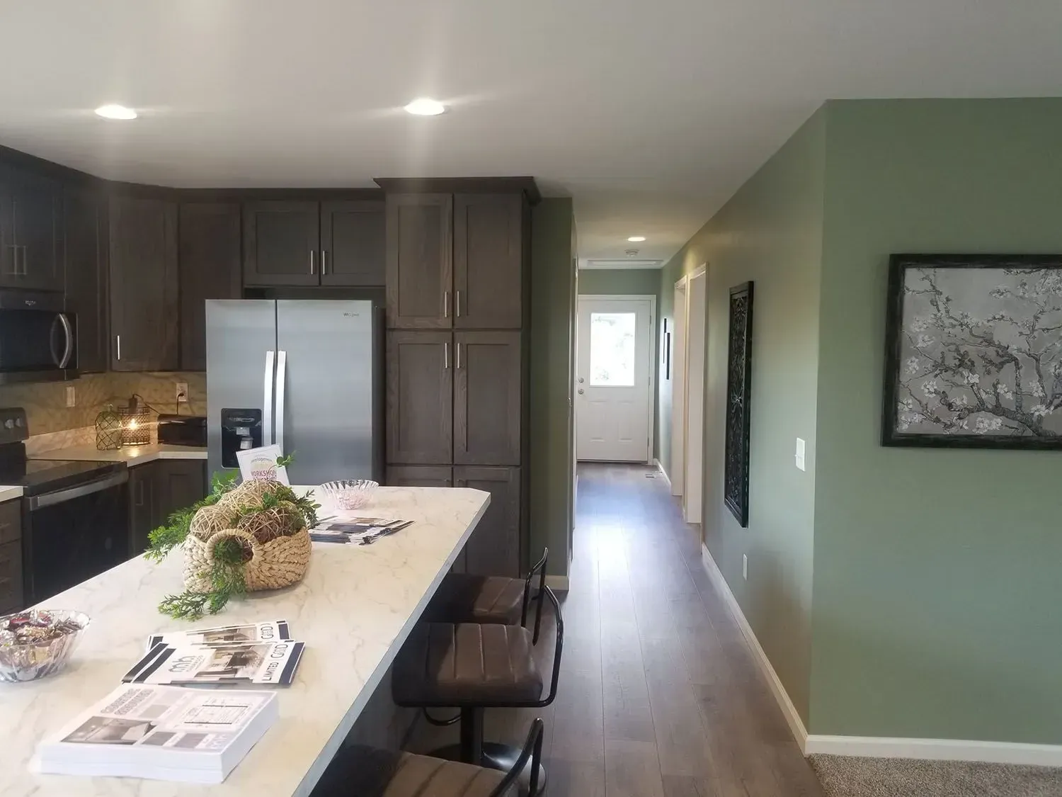Kitchen with dark cabinets, stainless steel refrigerator, and a hallway with sage green walls.