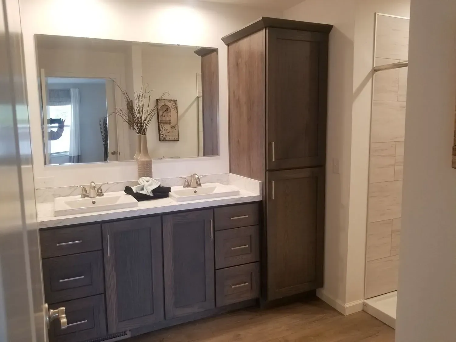 Bathroom with double sink vanity, tall storage cabinet, and shower. Dark gray cabinets, light countertops, and wood-look flooring.