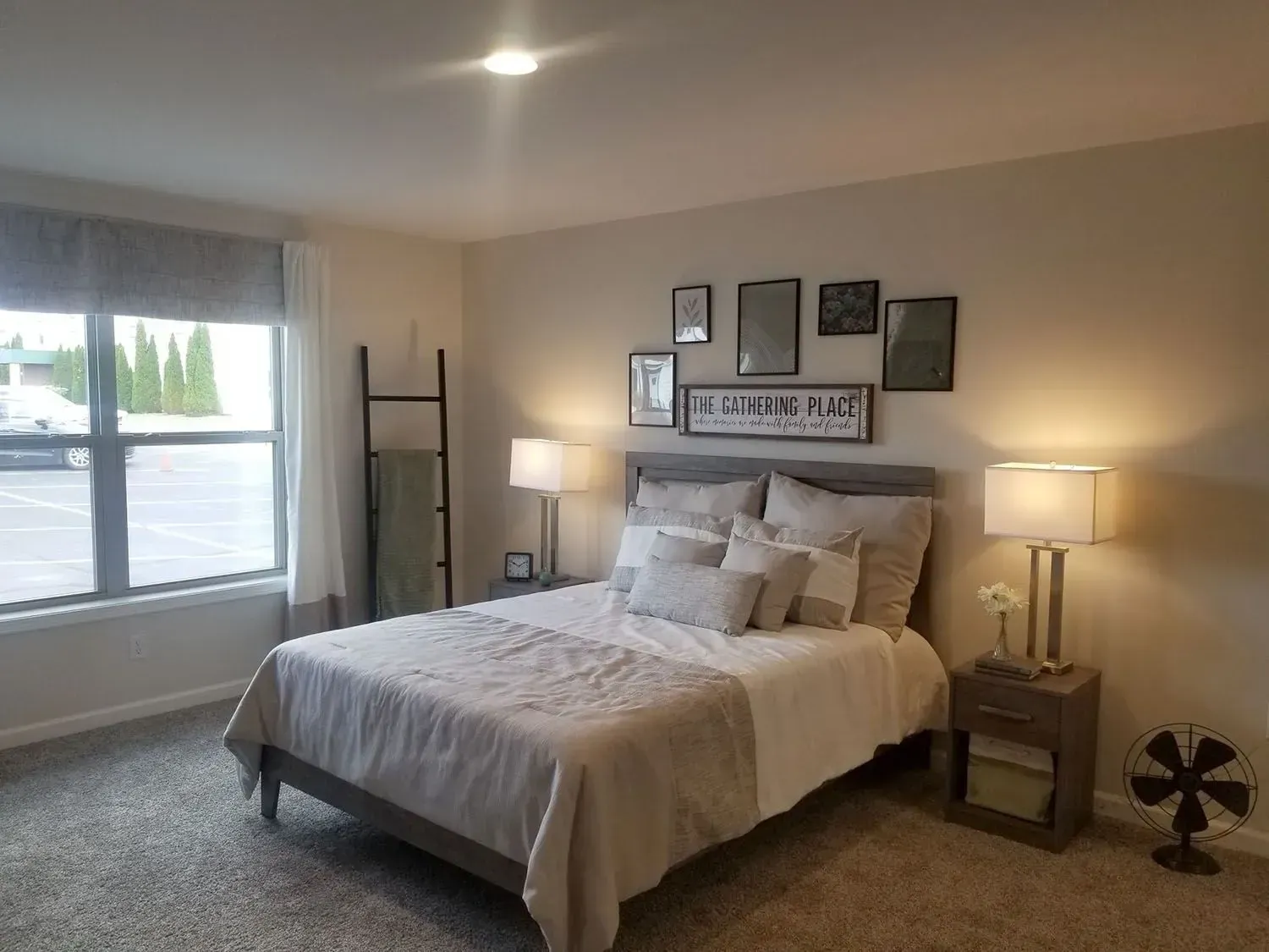 Bedroom with bed, nightstands, wall art, window, and a small fan. Beige and gray color scheme.