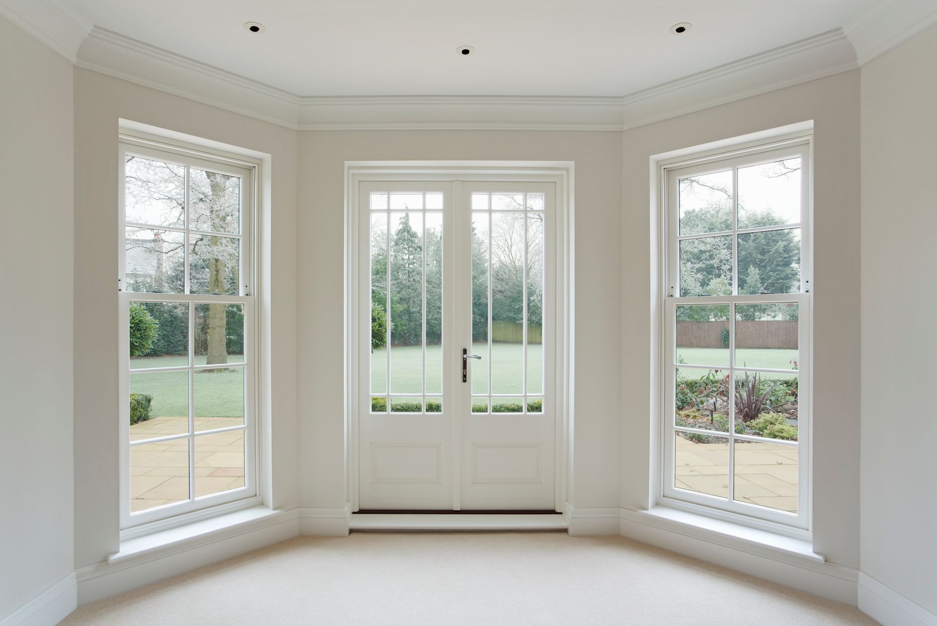 Bright empty bay room with three large windows and French doors overlooking a green yard