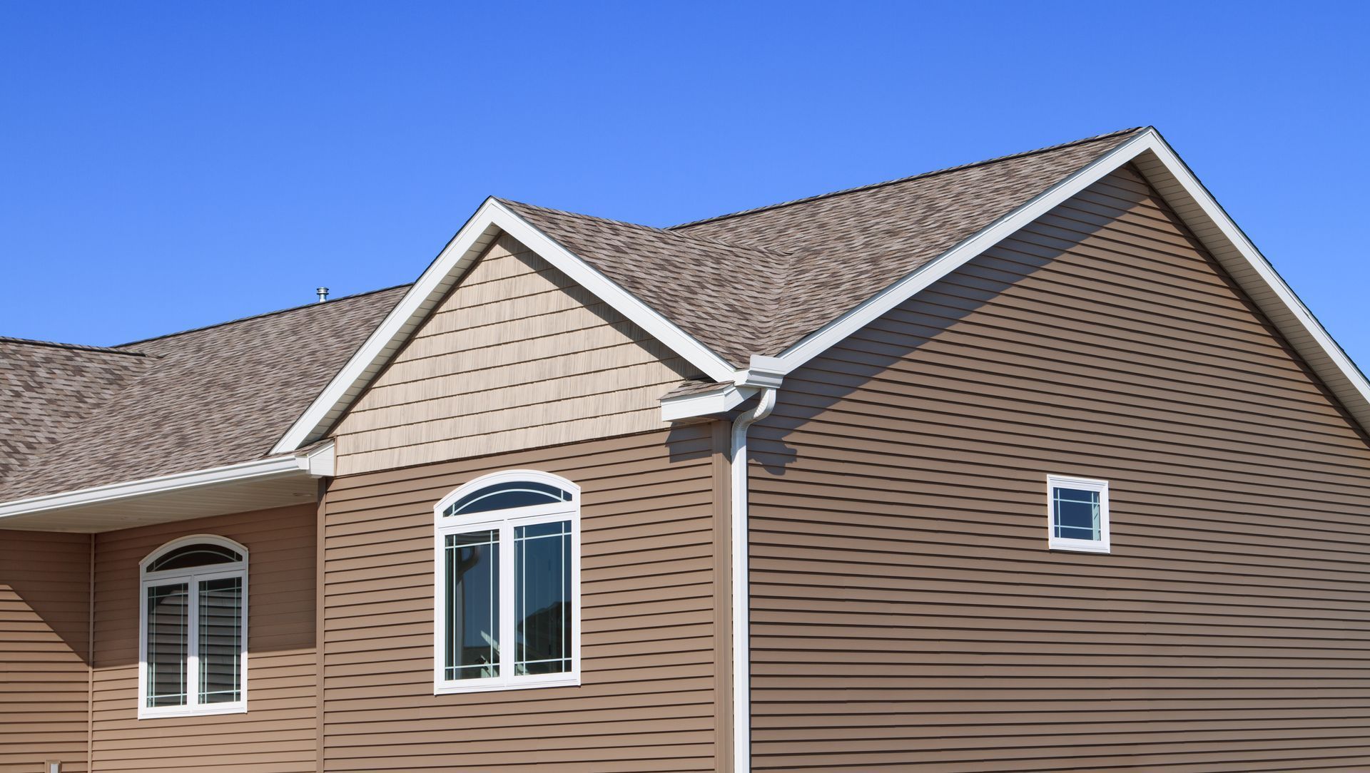 Suburban house roof with tan siding and white-trimmed windows against a blue sky