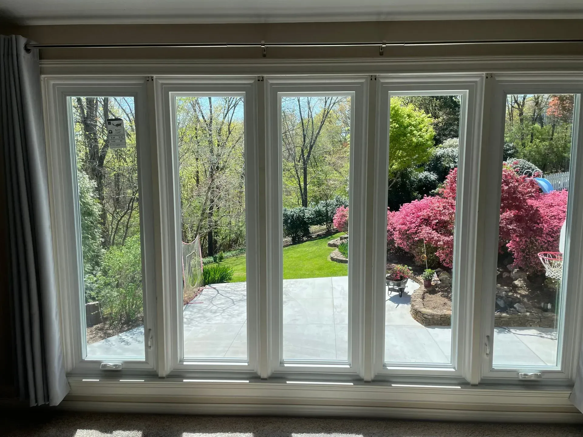 View through white-framed windows to a sunny garden with trees, pink bushes, and a stone patio