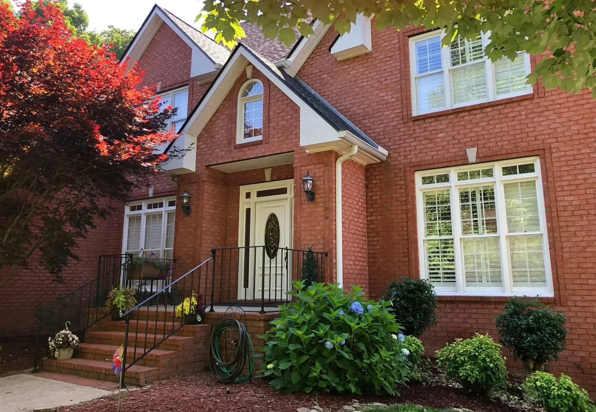 Brick house with white trim, front porch, and manicured shrubs beside a red-leafed tree.