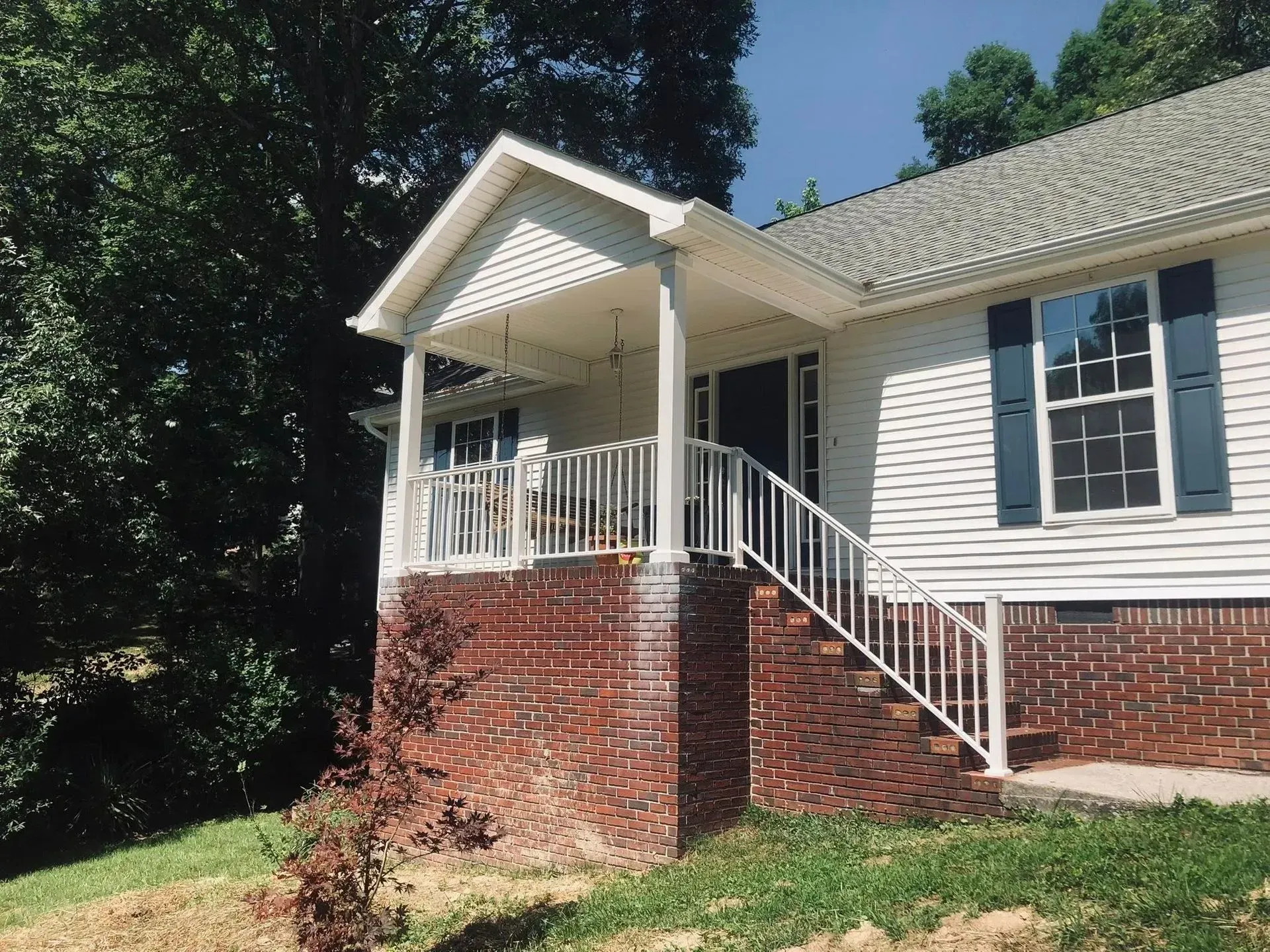 White house porch with brick foundation and white railings beside trees on a sunny day