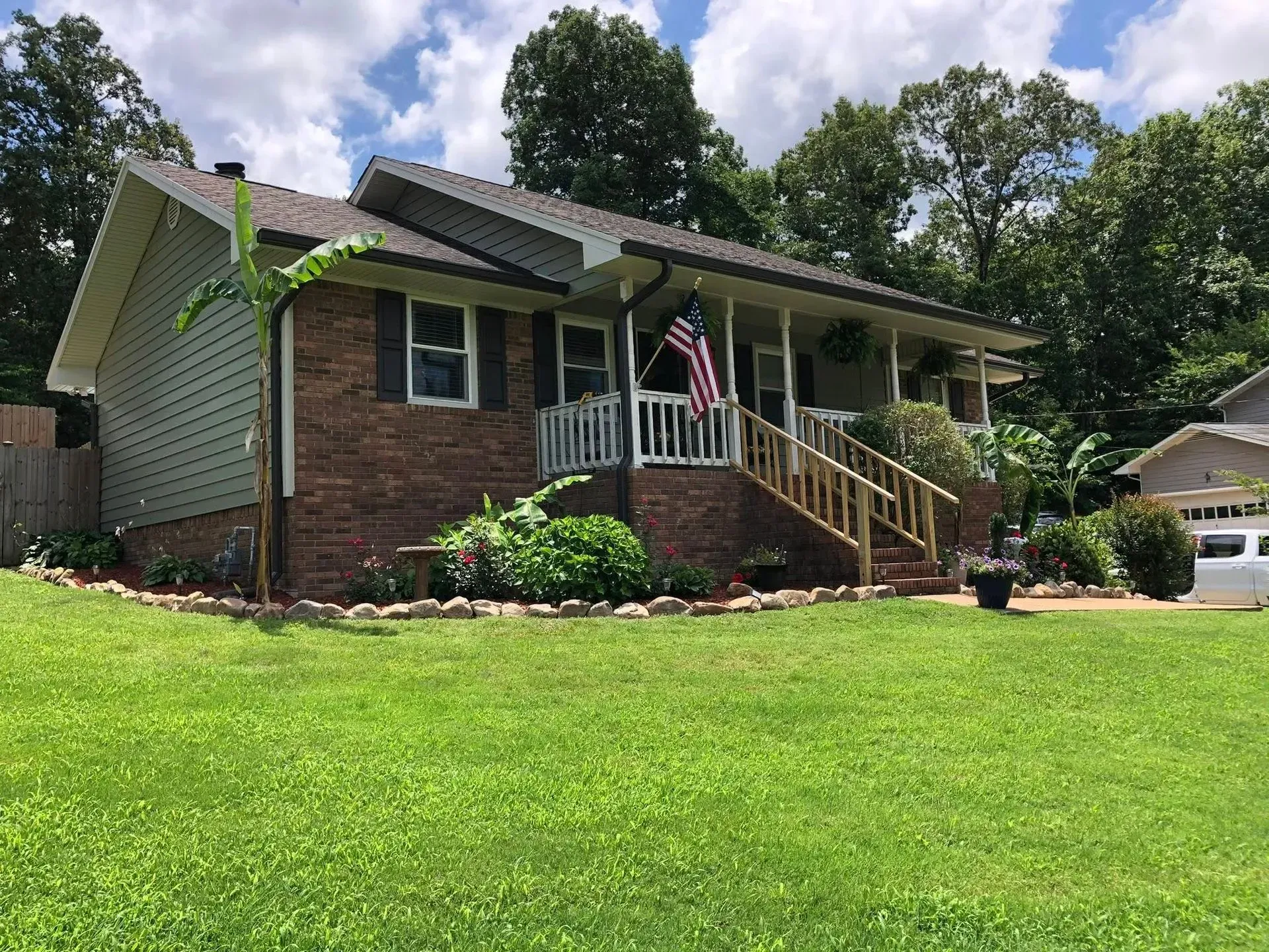 Suburban brick house with front porch, lawn, and an American flag under a partly cloudy sky