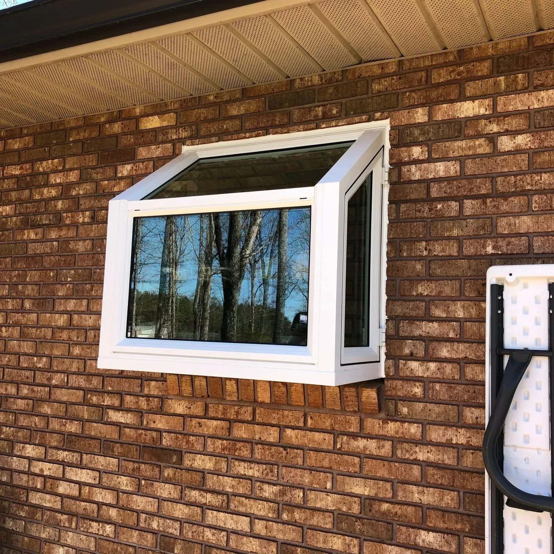 White awning window open on a brick house exterior, reflecting trees and sky.