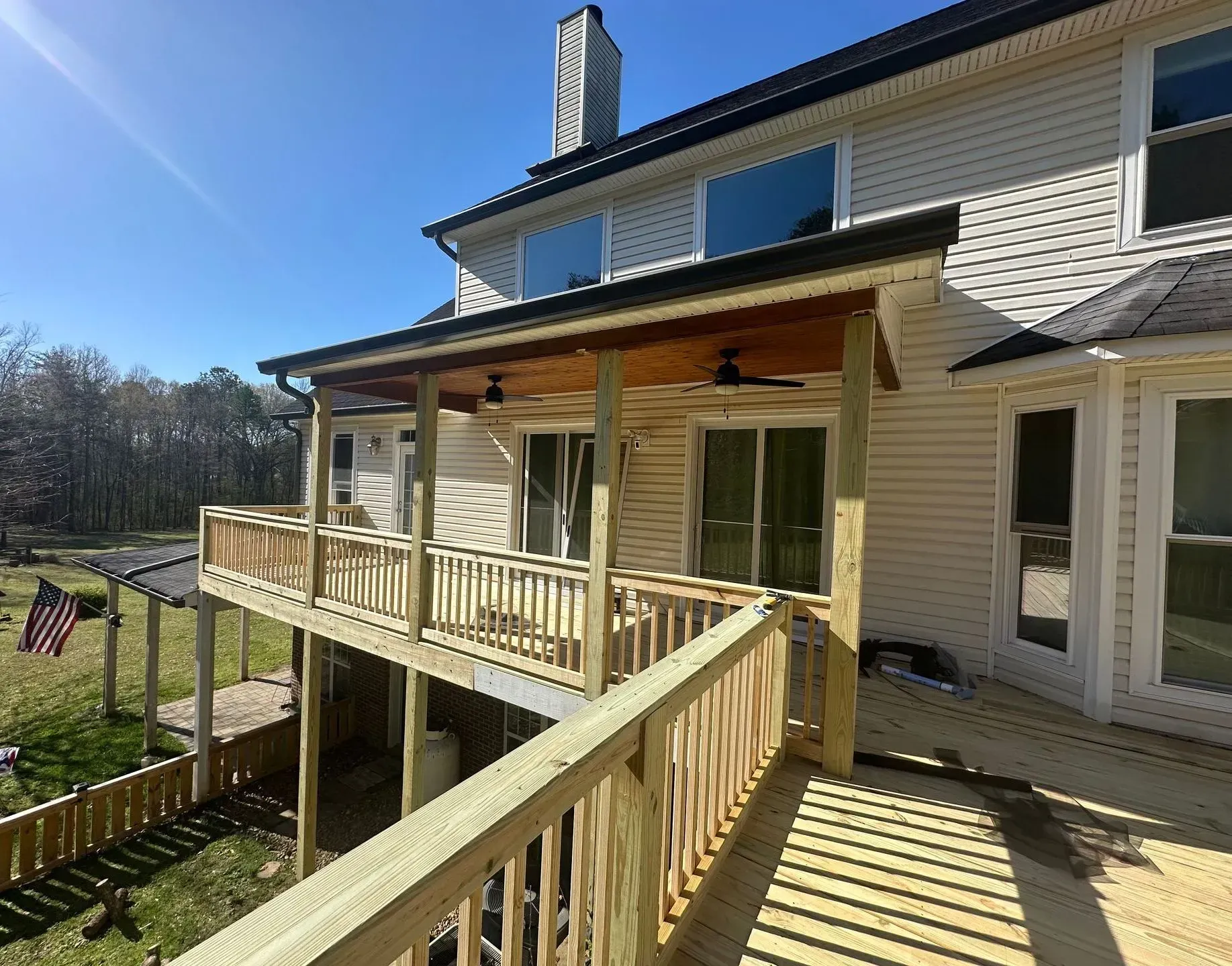 Beige house with a wooden wraparound porch and ramp in bright sunlight, viewed from the side.