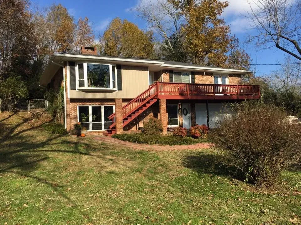 Two-story house with red deck on a grassy hillside, framed by autumn trees and blue sky
