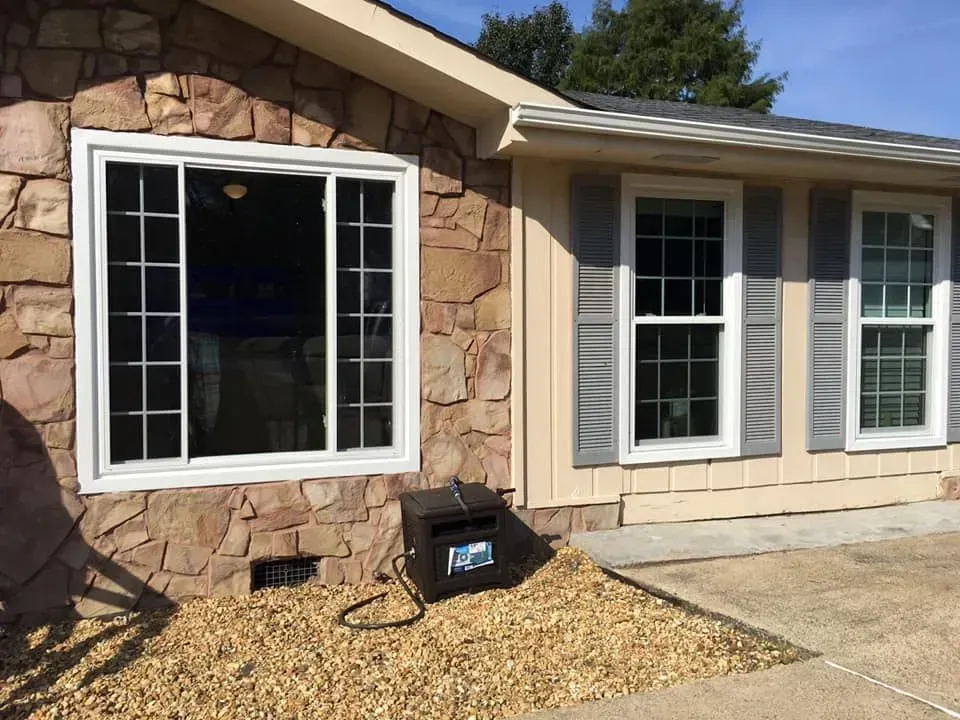 House exterior with a large white window, stone siding, and a small black unit on a gravel bed by the wall