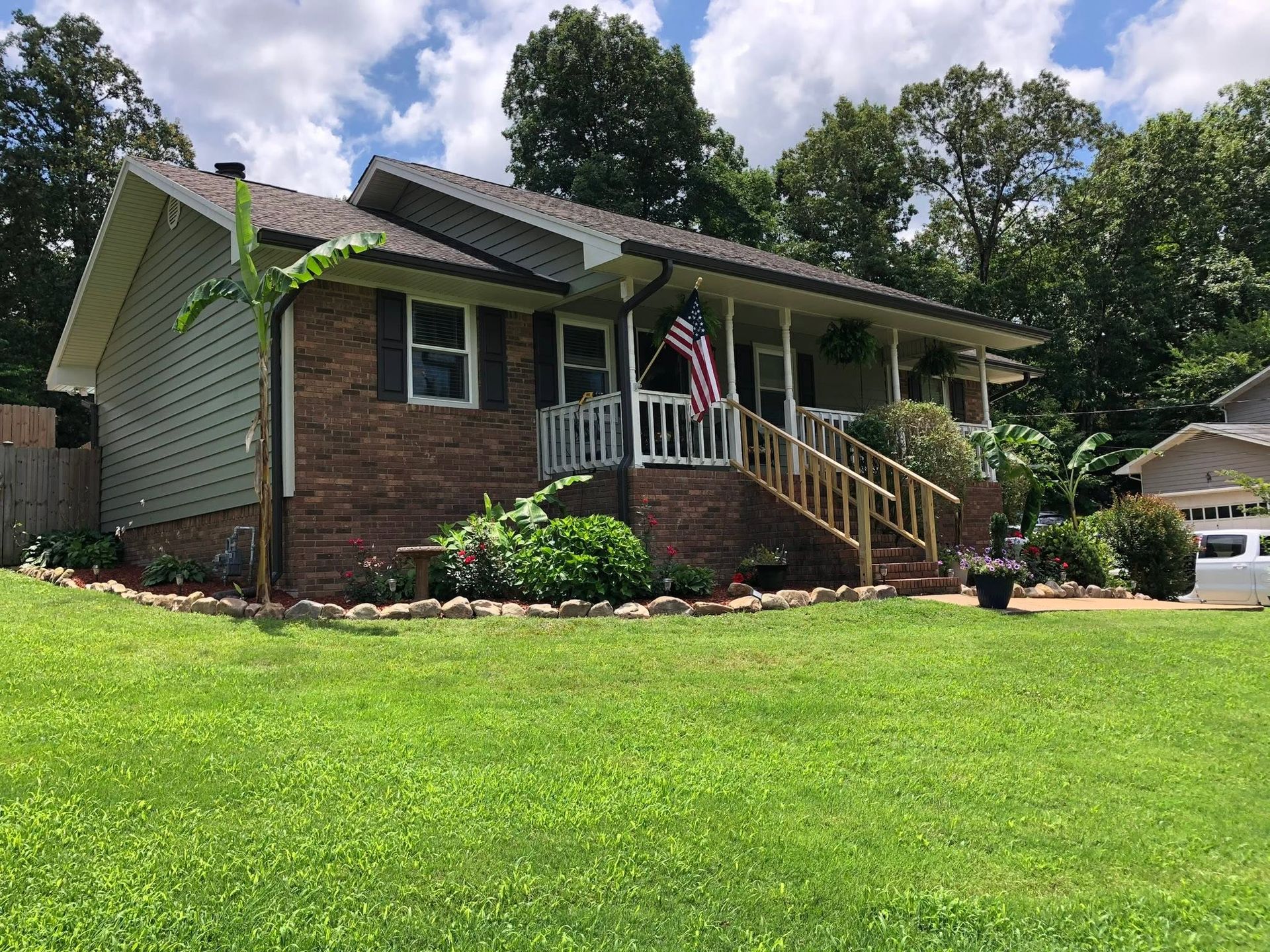 Suburban house with brick front, porch, American flag, and a green lawn under a partly cloudy sky