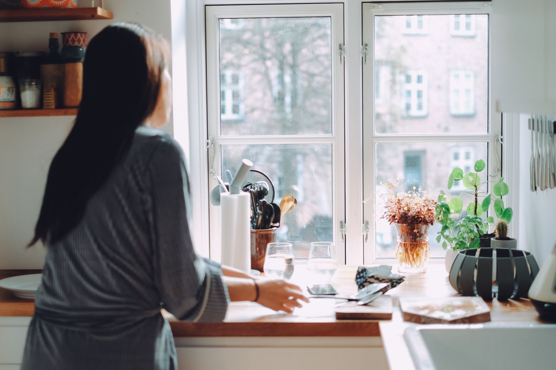 Person standing at a kitchen counter by a bright window, with plants and flowers on the sill.