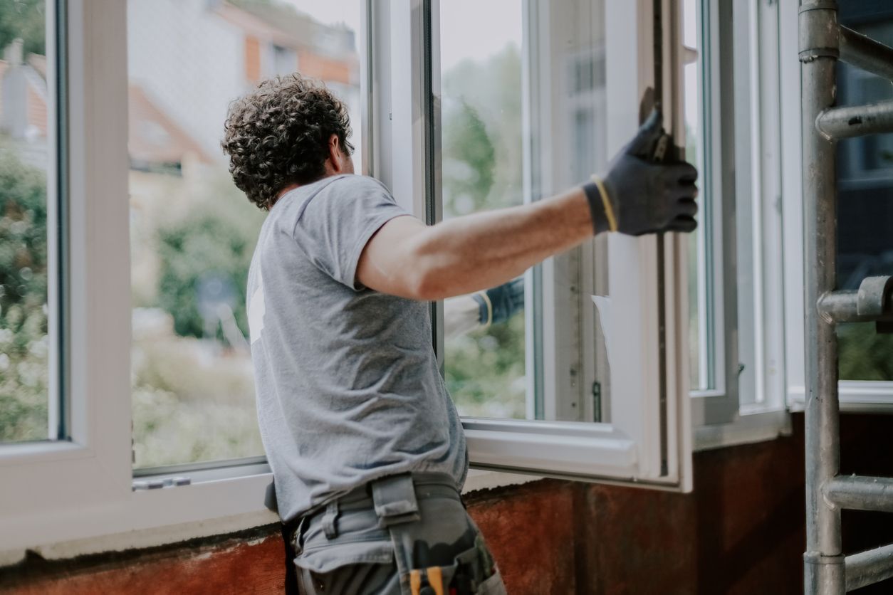 Worker installing a white window frame beside an open window, wearing gloves and a tool belt.