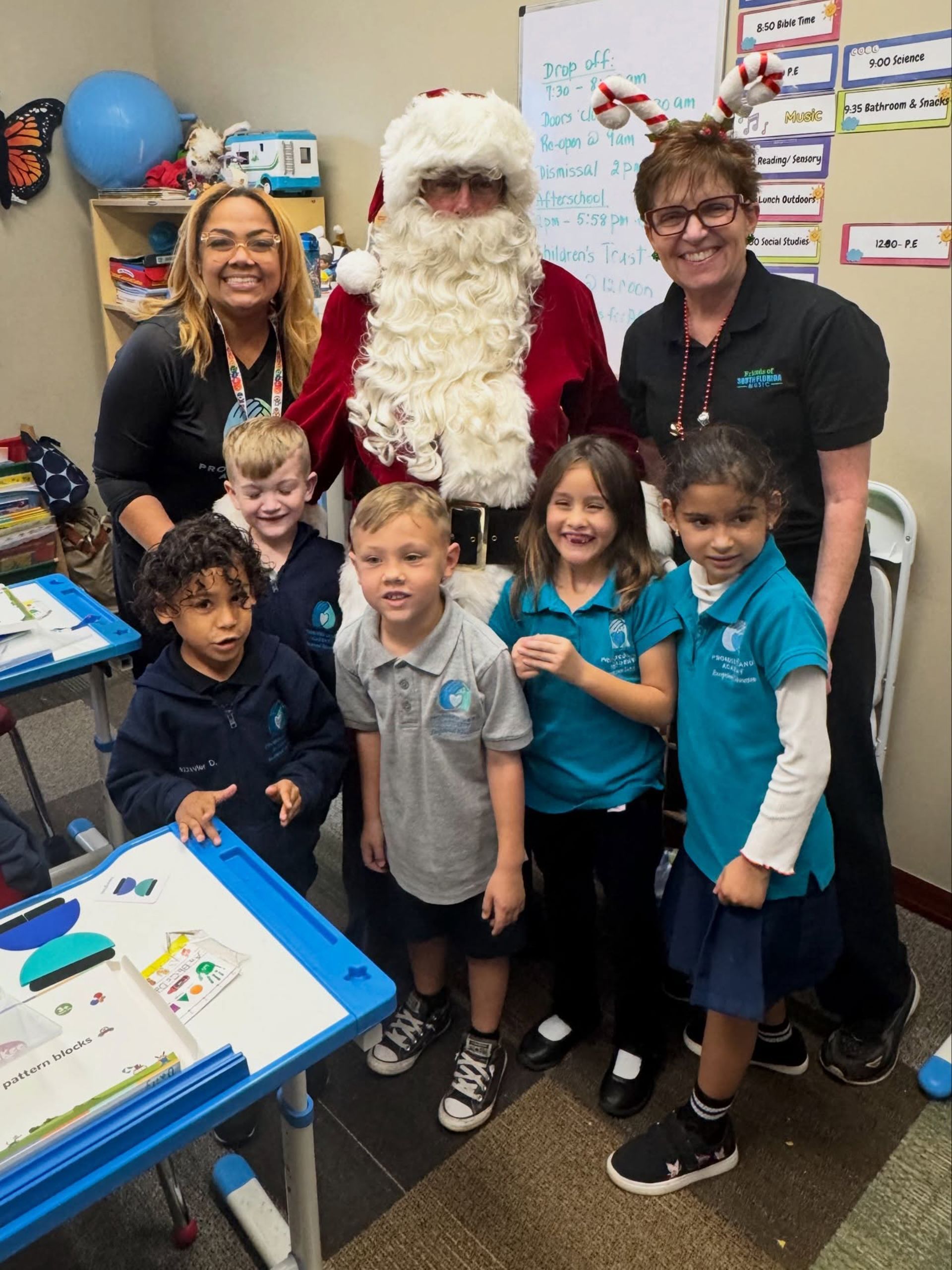 Santa posing with smiling children and two adults in a classroom during a holiday celebration