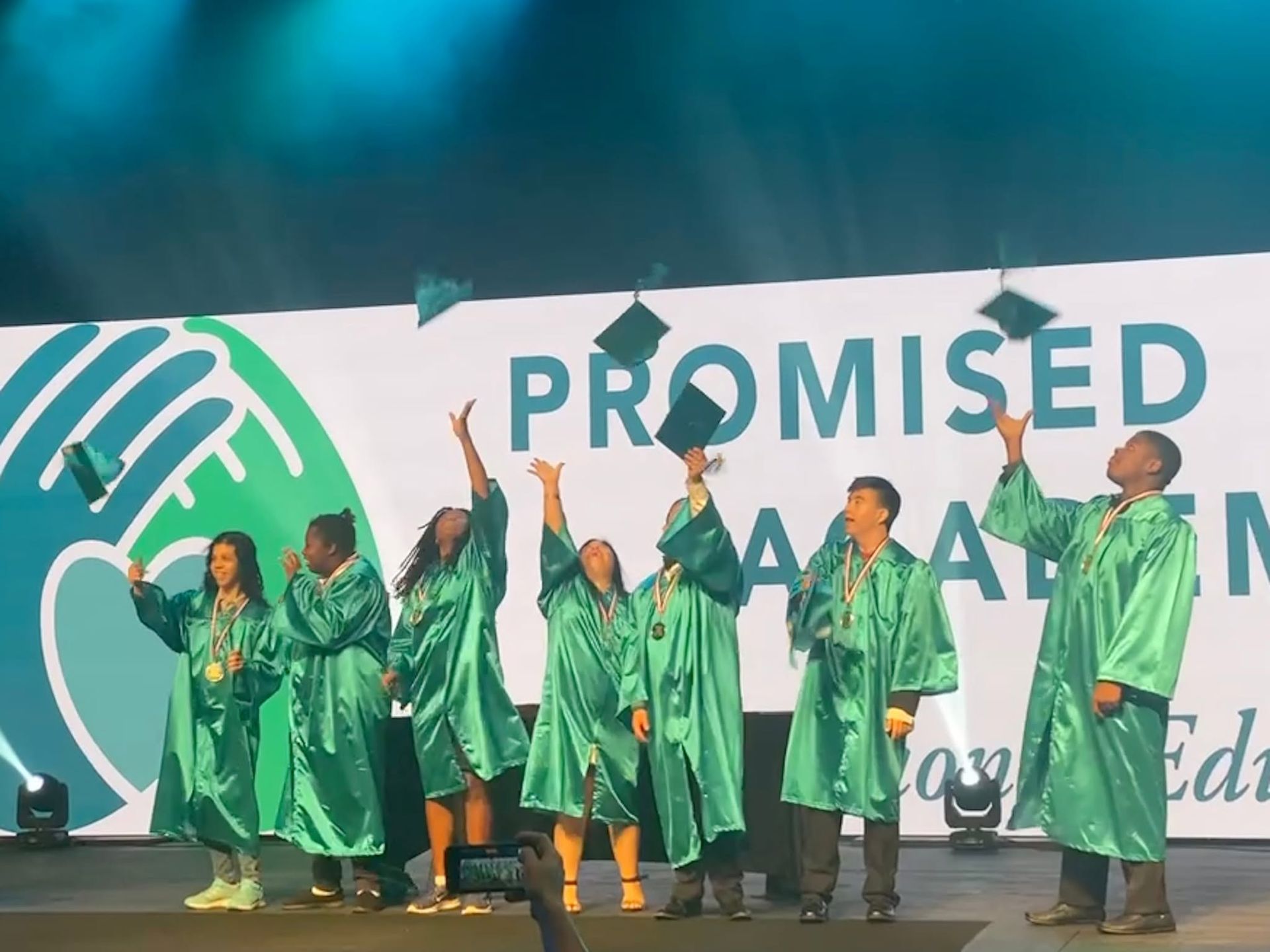 Performers in green robes onstage holding hands beneath a “PROMISED” banner