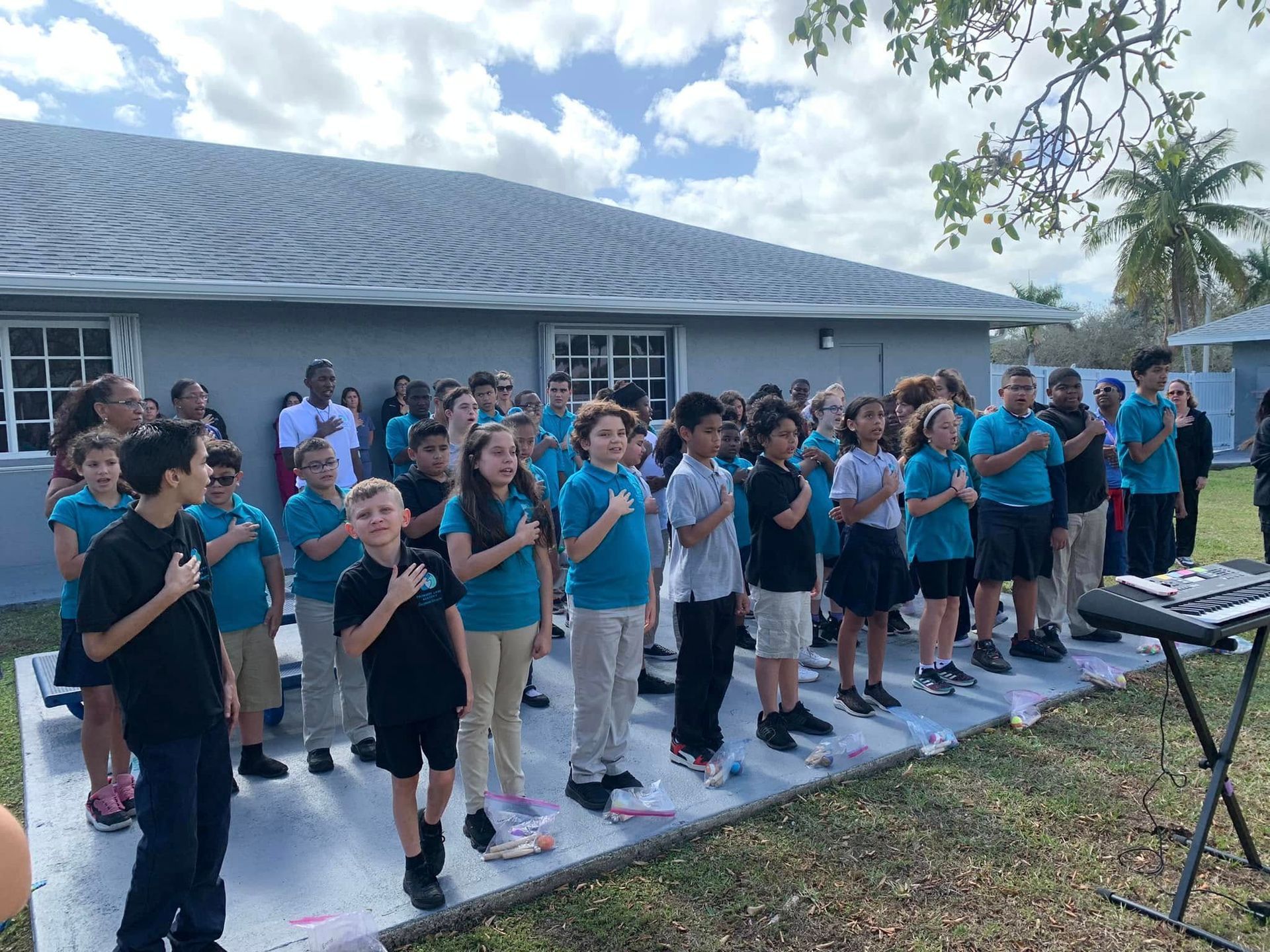 Large group standing outdoors in front of a building, singing or reciting together near a keyboard stand