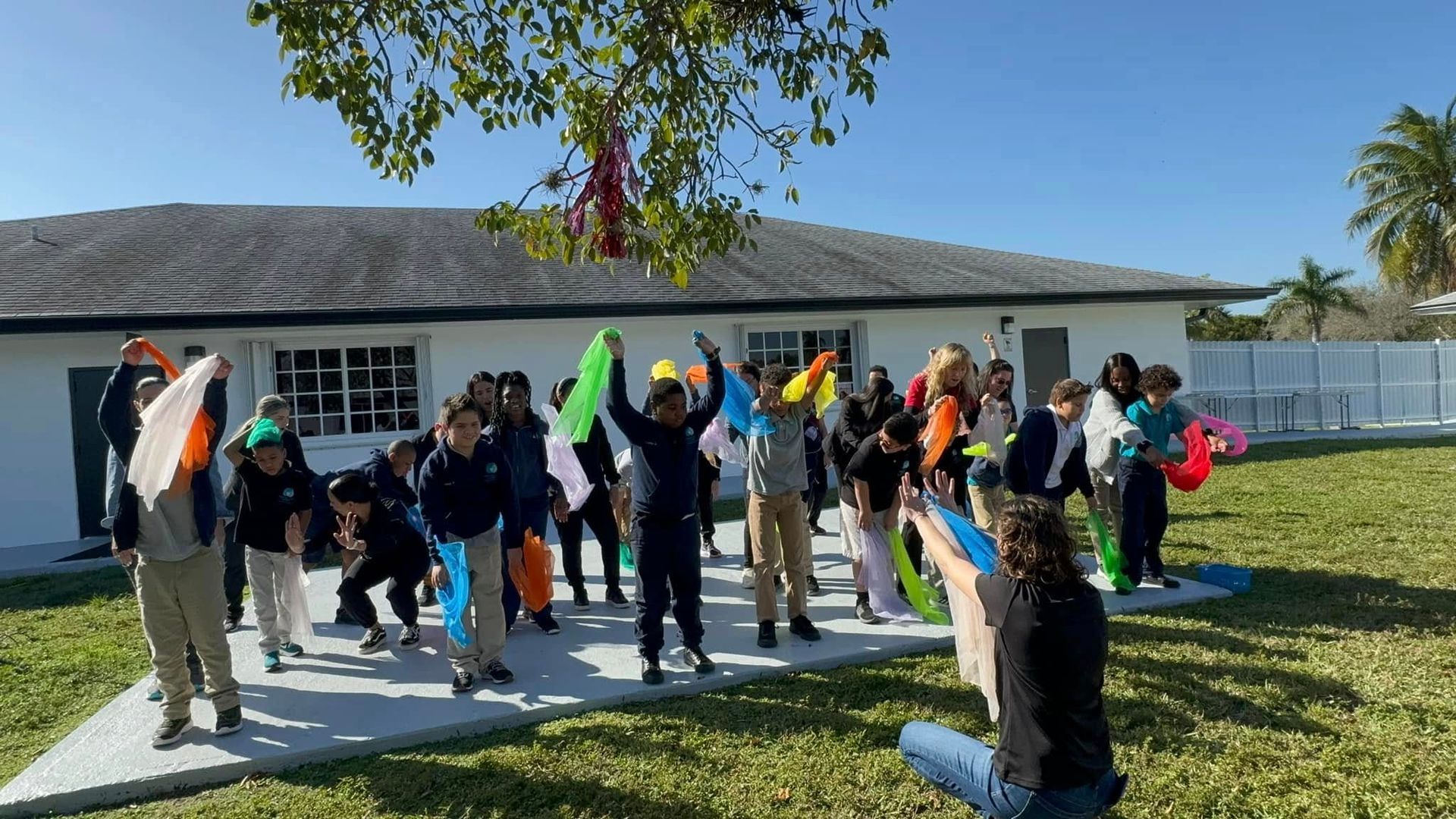 Group of people outdoors holding colorful shoes in front of a white building and palm tree