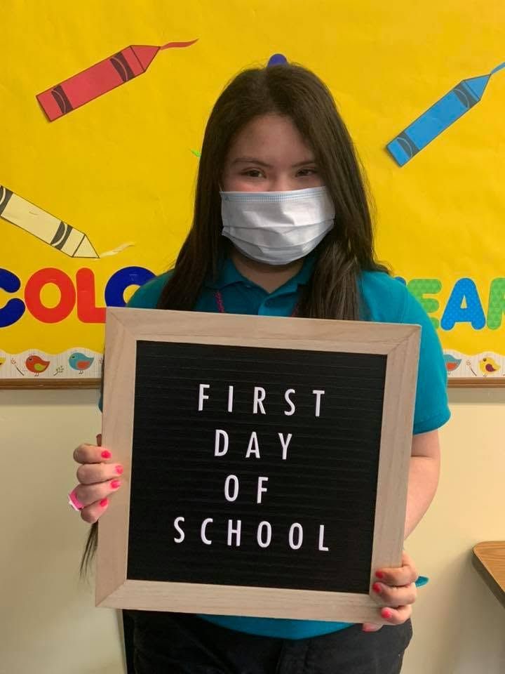 Student holding a sign that says “FIRST DAY OF SCHOOL” in a colorful classroom.
