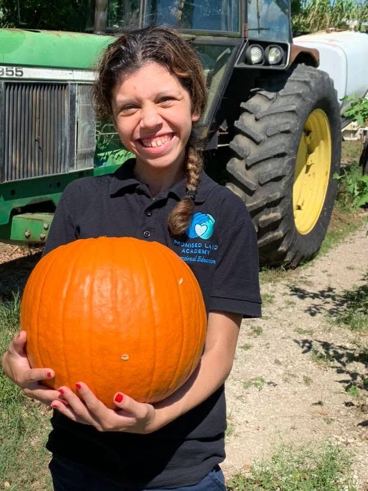 Smiling person holding a large orange pumpkin in front of a green tractor