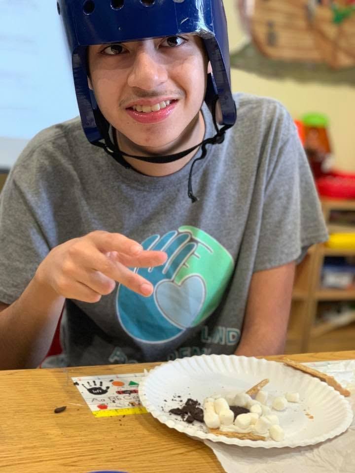 Child wearing a blue helmet smiles at a table with a plate of food.