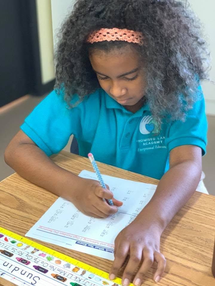 Student in a blue polo shirt writes on a worksheet at a wooden desk in a classroom.