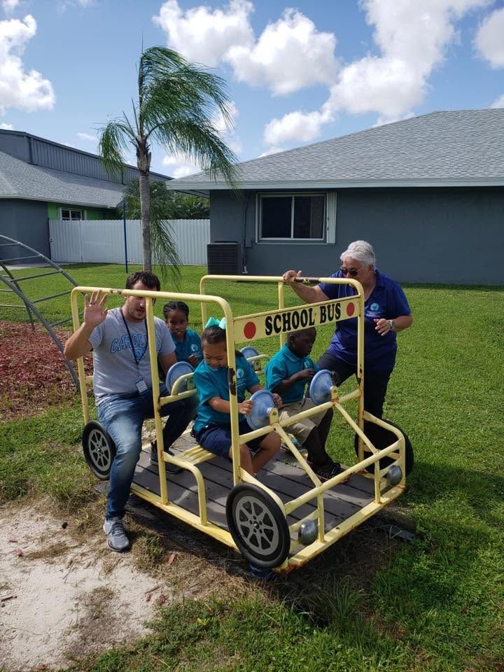 Three boys on a yellow pedal cart with an adult pushing in a driveway near a house