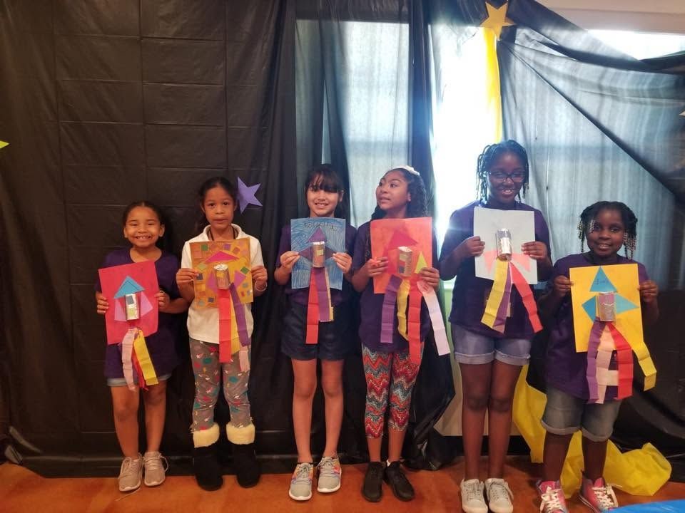Children holding colorful paper crafts on a stage, standing in front of black curtains and windows.