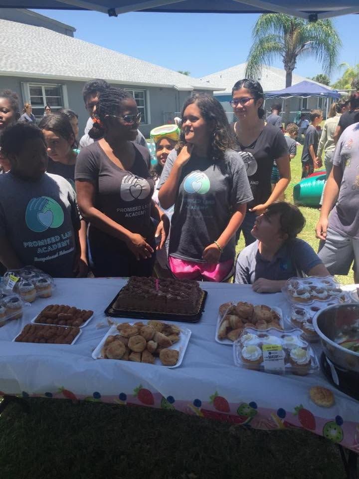People chatting at an outdoor bake sale table with assorted pastries and desserts under a sunny canopy