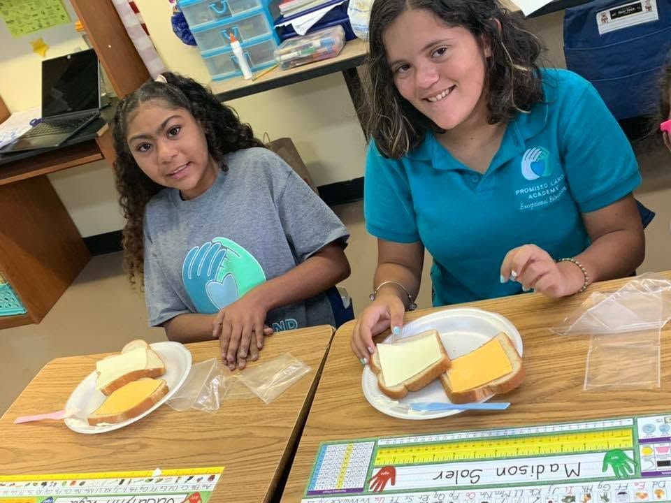 Two students smiling at a table with sliced bread and cheese in a classroom setting