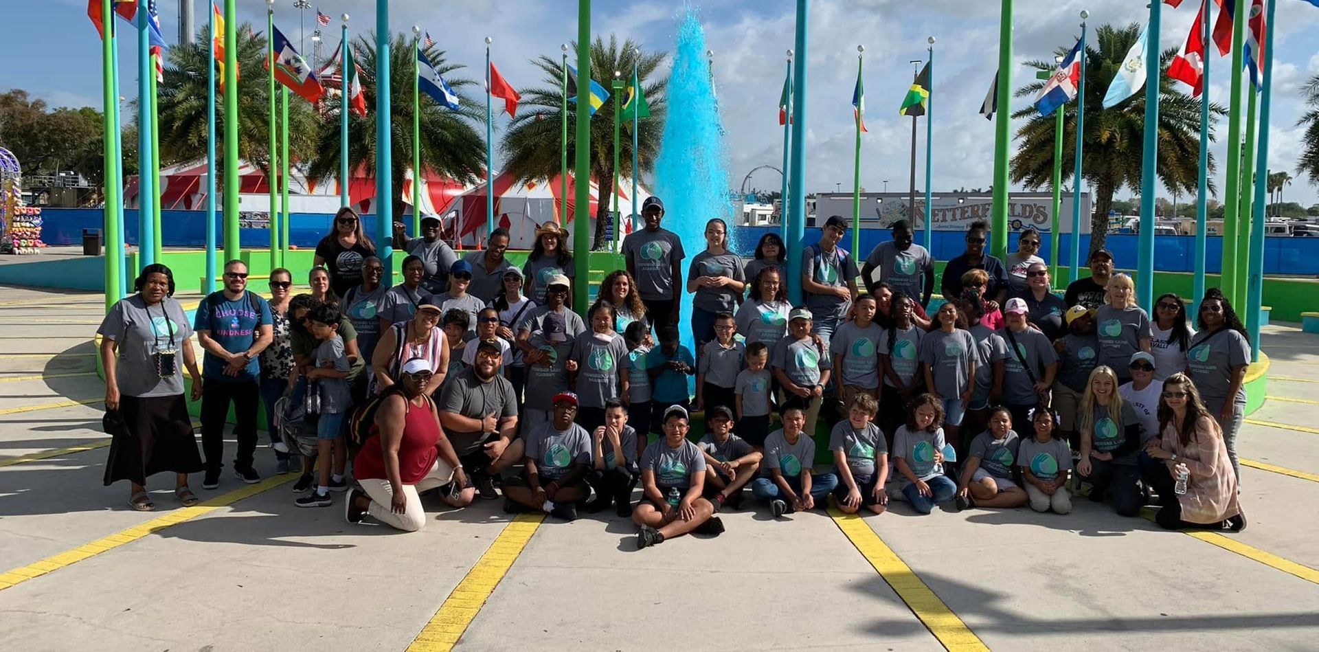 Large group posing outdoors under flags in front of a blue building, with yellow road markings in foreground