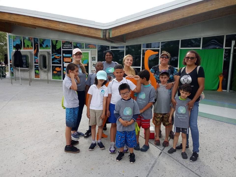Group of children and two adults standing outdoors in front of a school building, smiling at the camera.