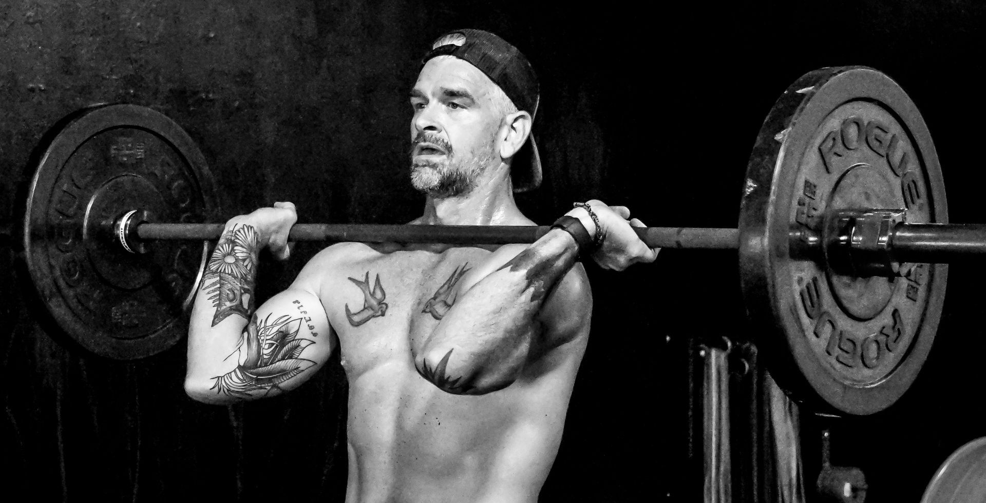 Man lifting barbell, indoors. He's tattooed, shirtless, wearing a cap, with a focused expression. Black and white.