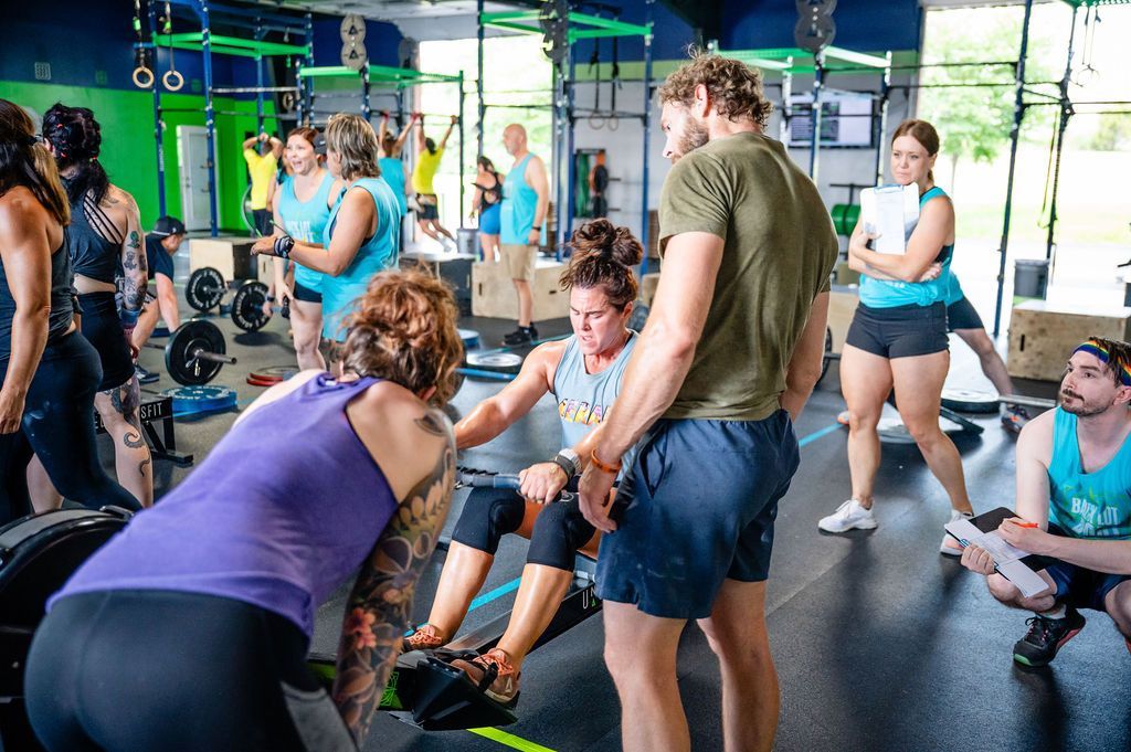 Group of people exercising in a gym, gathered around a trainer with kettlebells and rowing machines.