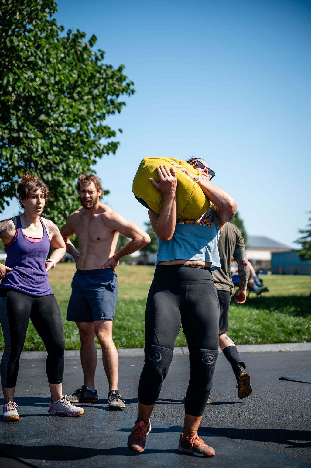 Athlete in blue shirt and black leggings lifting a yellow sandbag outdoors, with spectators nearby