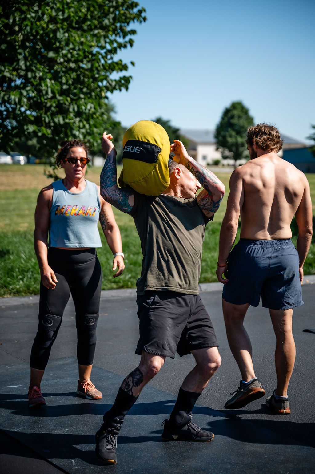 Three people exercising outdoors on a sunny road, one lifting a yellow bag over their head.