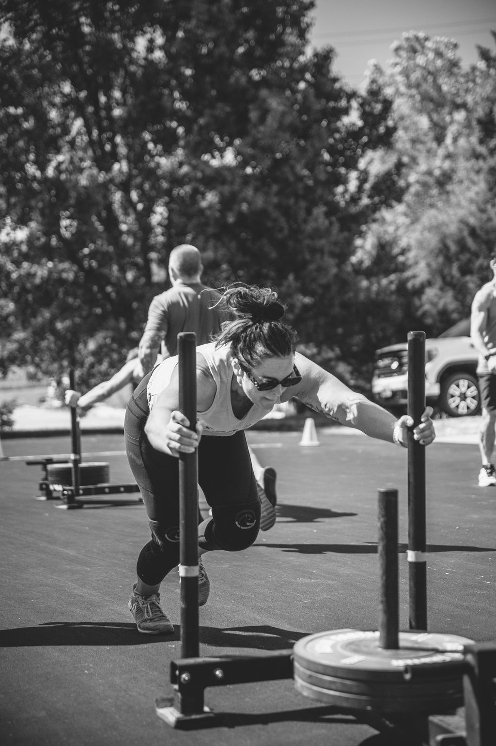Person pushing a weighted sled outdoors on a paved lot, with trees and a parked car in the background.