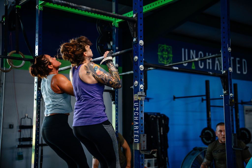 Two women doing pull-ups in a gym with blue walls and green rigging