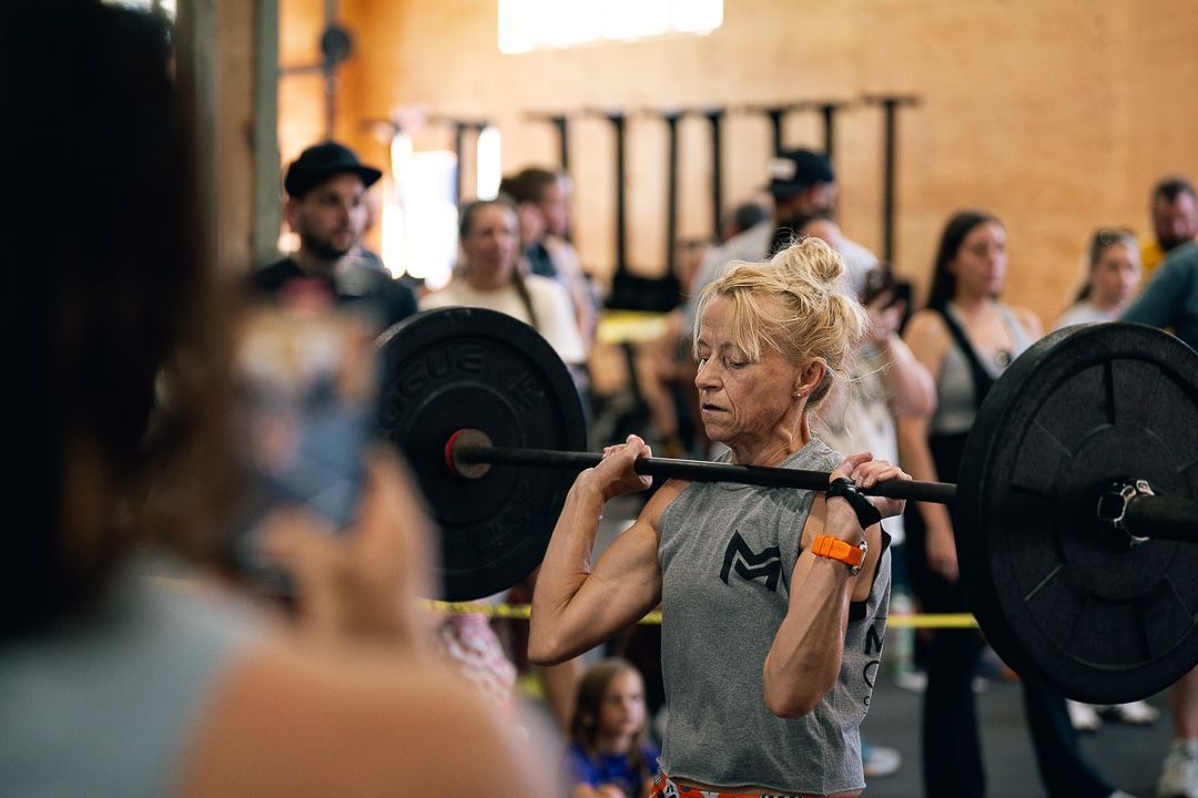 Person lifting a barbell overhead in a gym, with people watching in the background.