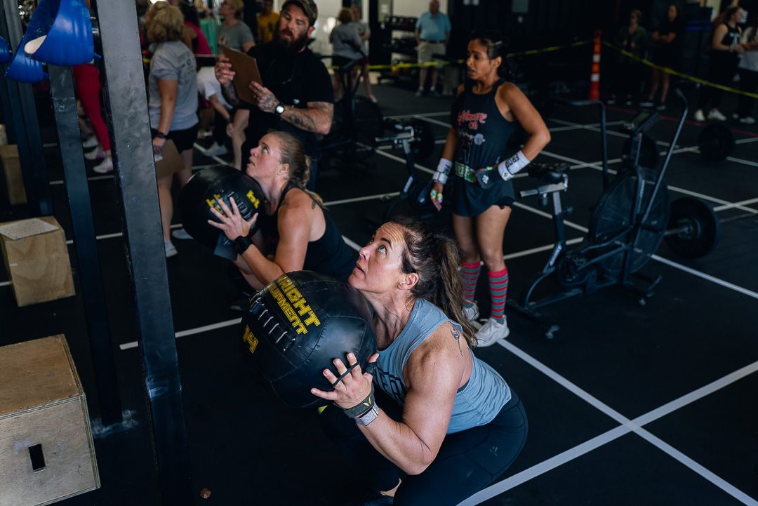People exercising with medicine balls in a gym, squatting on a marked floor.