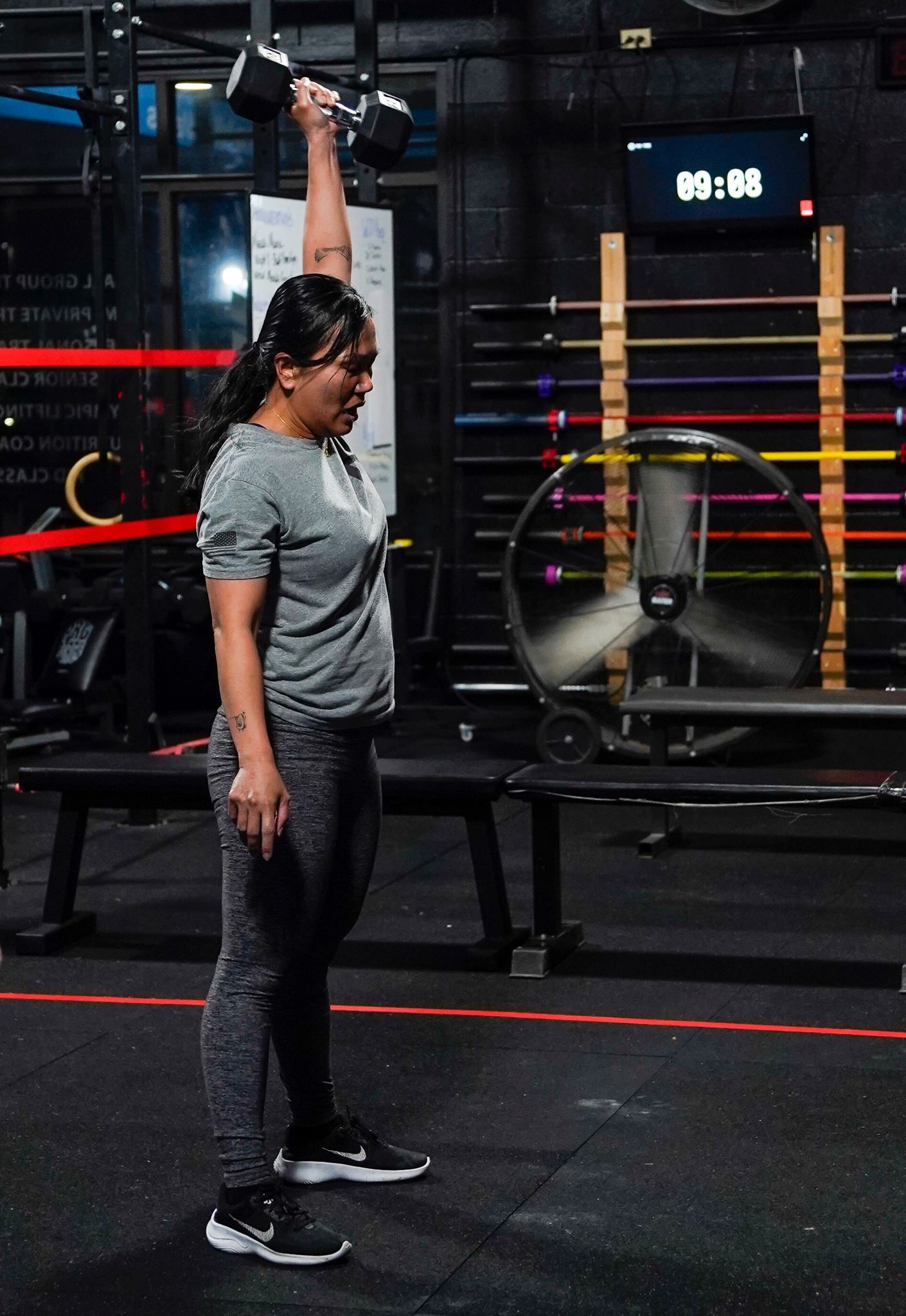 Woman in a gym holding a kettlebell overhead beside a bench and rack of colored bars.