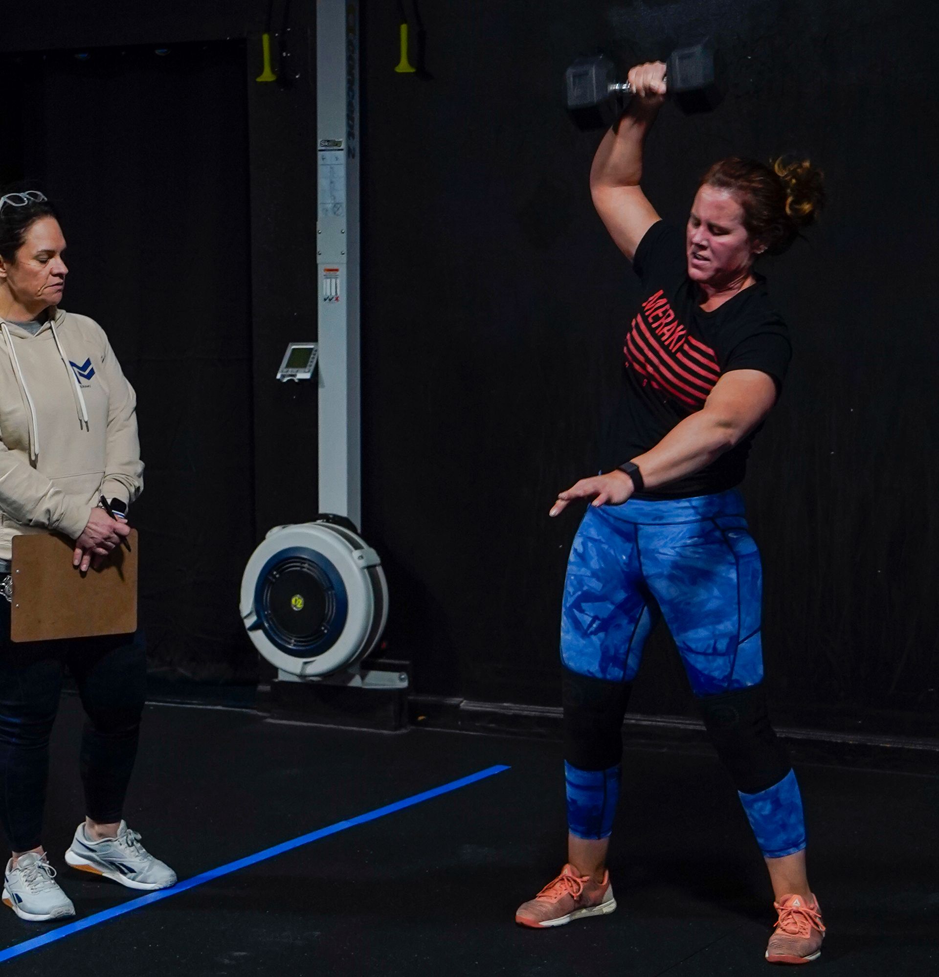 Two women in a gym; one lifts a dumbbell overhead while the other watches with a clipboard.