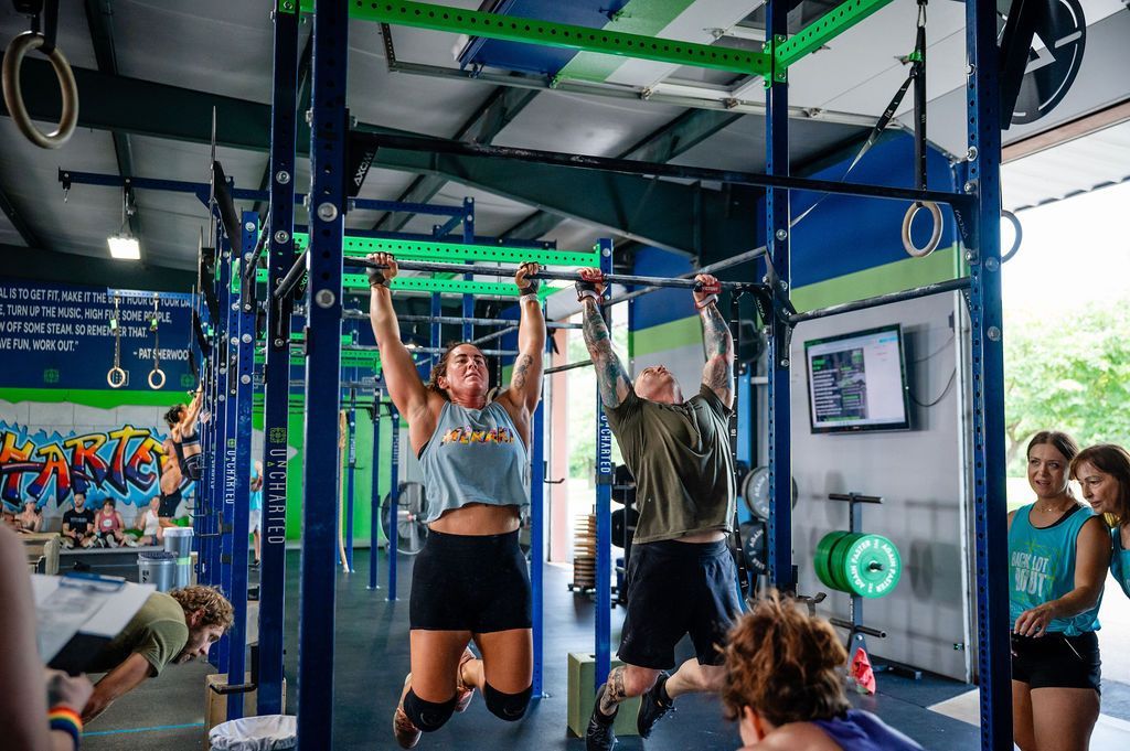 Two people doing pull-ups in a blue gym, with onlookers and colorful wall art nearby