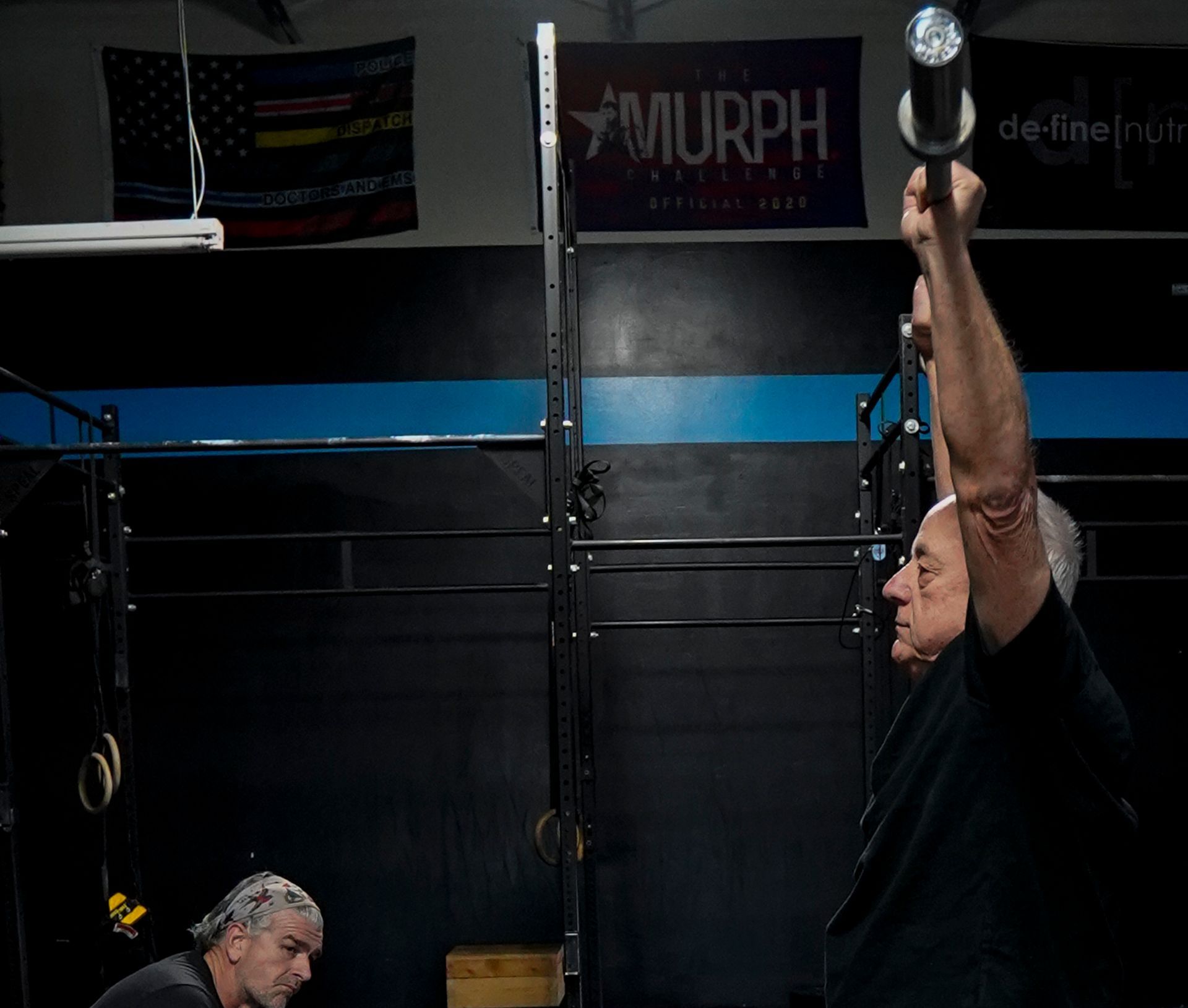 Gym athlete hanging from a pull-up bar in a dark CrossFit gym while another person looks on.
