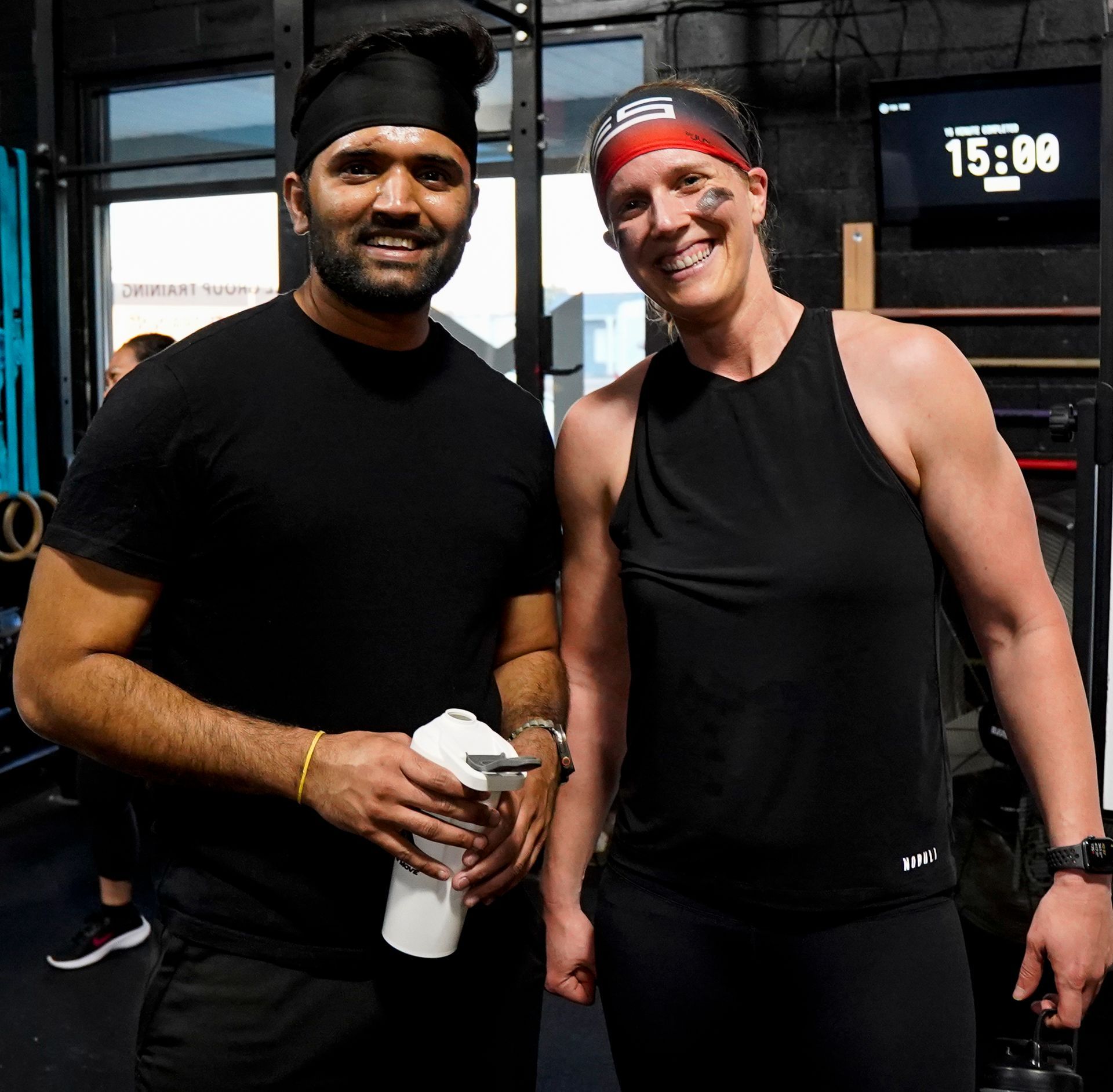 Two people in black gym clothes posing indoors, one holding a water bottle, with a digital clock behind them.