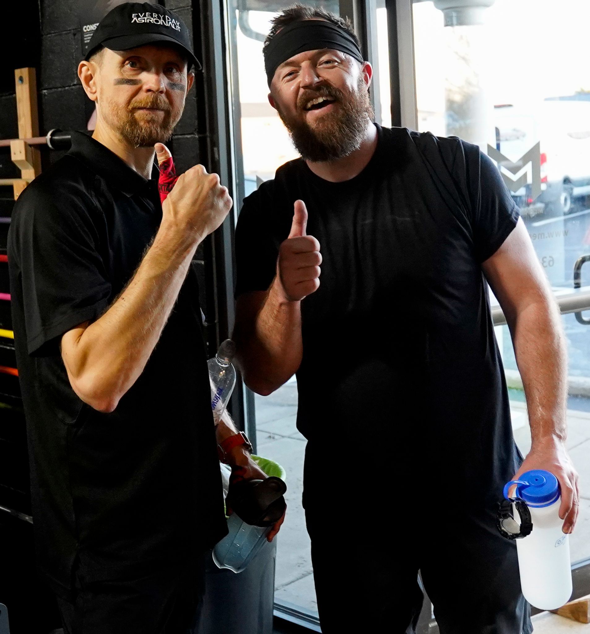 Two men in black shirts giving thumbs up in a gym, one holding a bottle