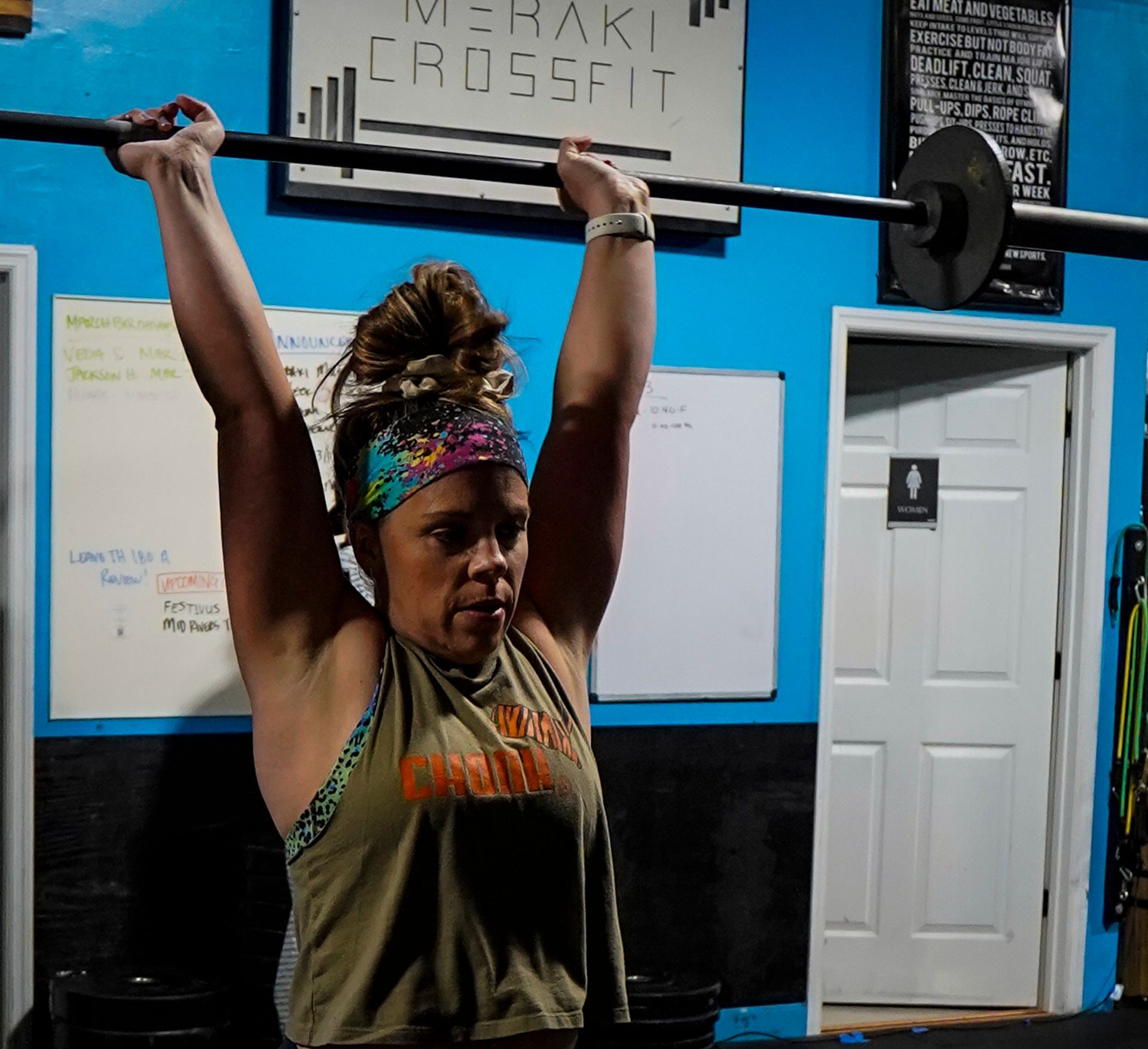 Woman lifting a barbell overhead in a gym, with blue walls, whiteboards, and a door behind her.